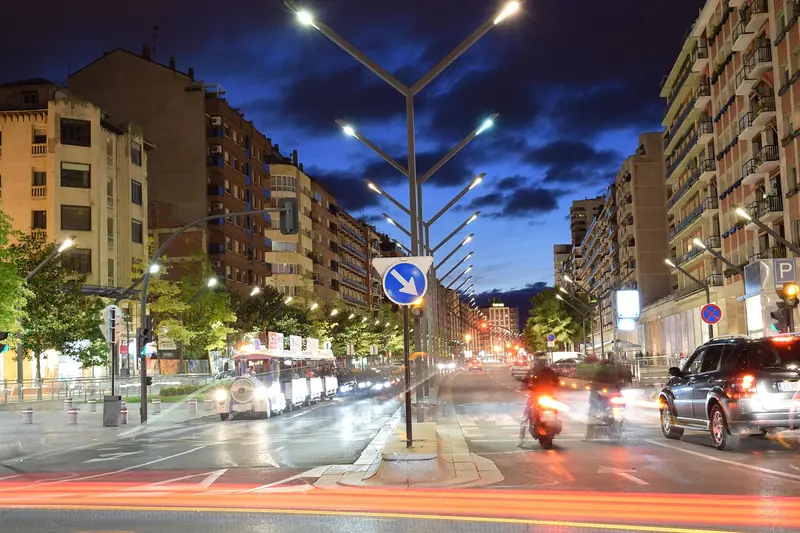 Avenida movimentada à noite, com motos e carros passando entre prédios residenciais
