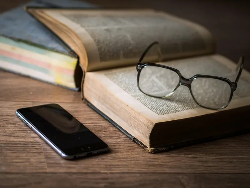 Livro aberto com óculos por cima e um celular ao lado, sobre uma mesa de madeira