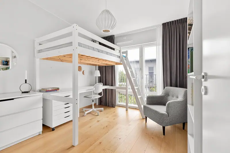 Quarto claro com cama alta, escrivaninha branca e poltrona perto da porta de vidro da varanda