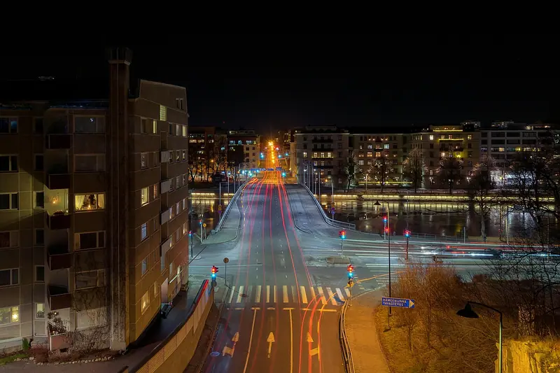 Avenida iluminada à noite entre prédios residenciais, com rastros de luz dos carros