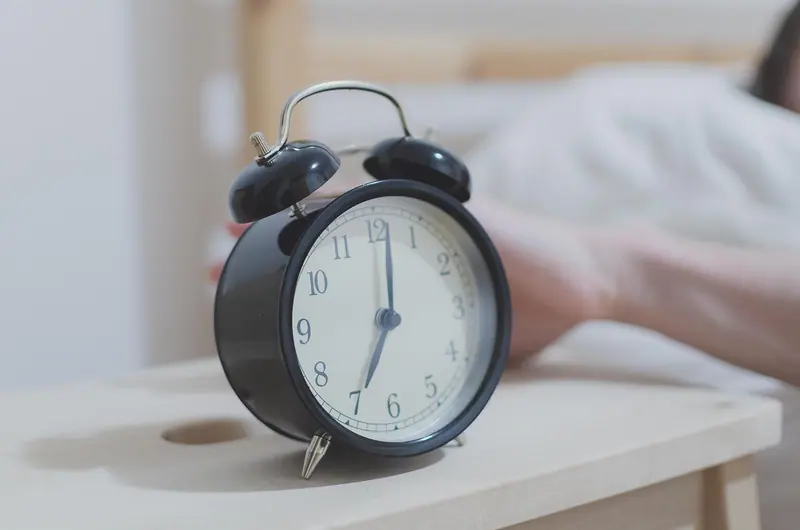 Mão saindo da cama em direção a um despertador preto sobre a mesa de cabeceira