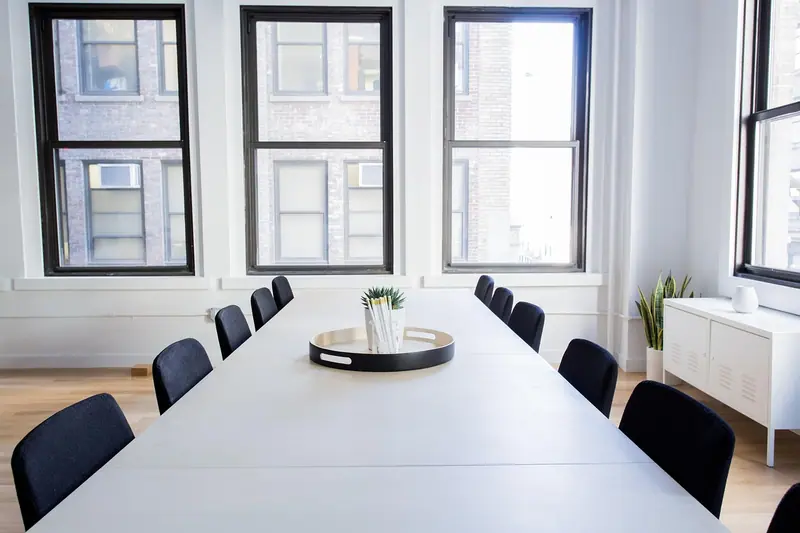 Sala de reunião clara com mesa longa branca, cadeiras pretas e janelas altas