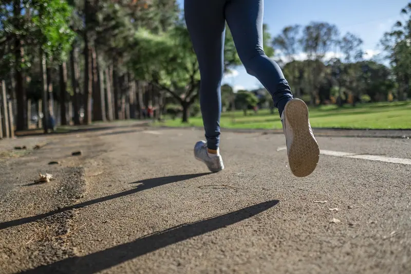 Pernas de uma pessoa correndo num caminho asfaltado de um parque arborizado
