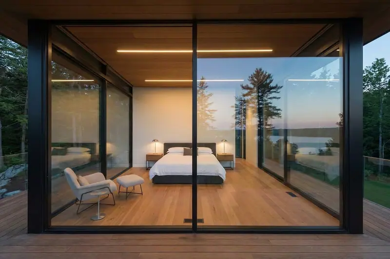 Quarto com paredes de vidro, cama arrumada e poltrona, aberto para árvores ao entardecer