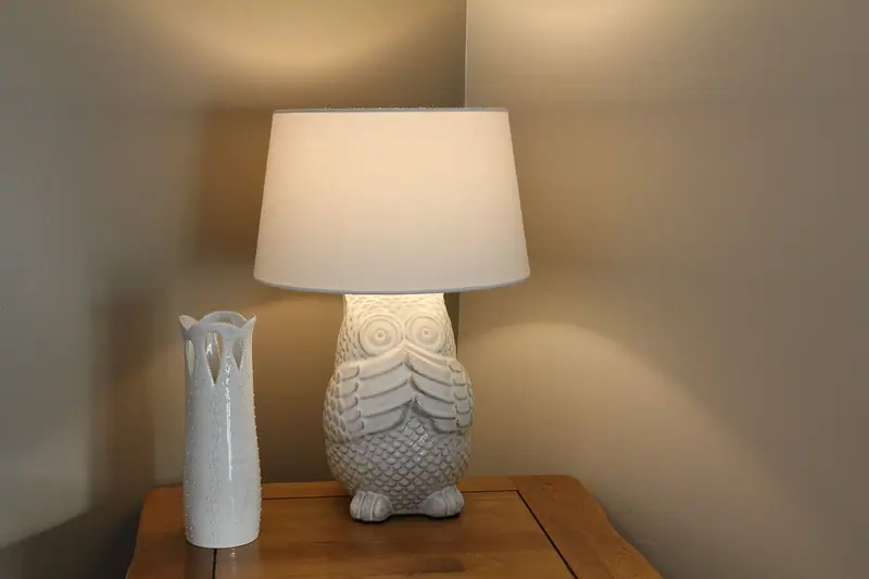 Abajur aceso sobre a mesa de cabeceira, ao lado de um vaso branco, num quarto à meia-luz