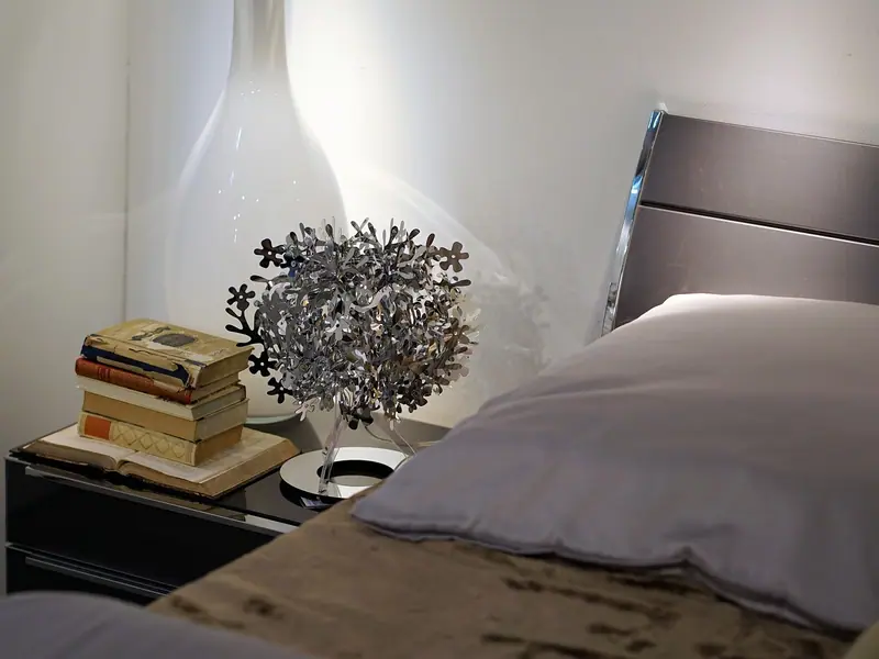 Mesa de cabeceira com livros empilhados e um arranjo de flores secas, ao lado de uma cama com lençóis cinza