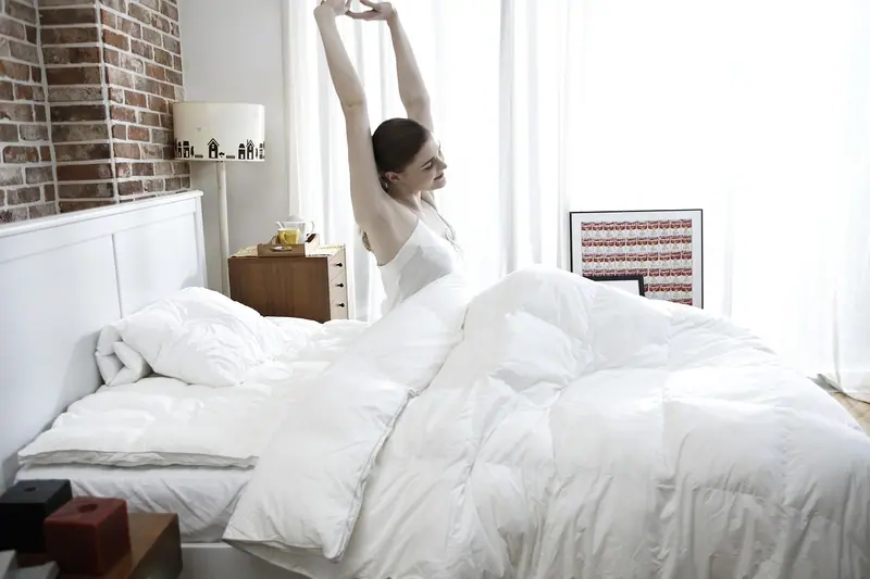 Mulher se espreguiçando ao lado da cama, num quarto claro com cortinas brancas