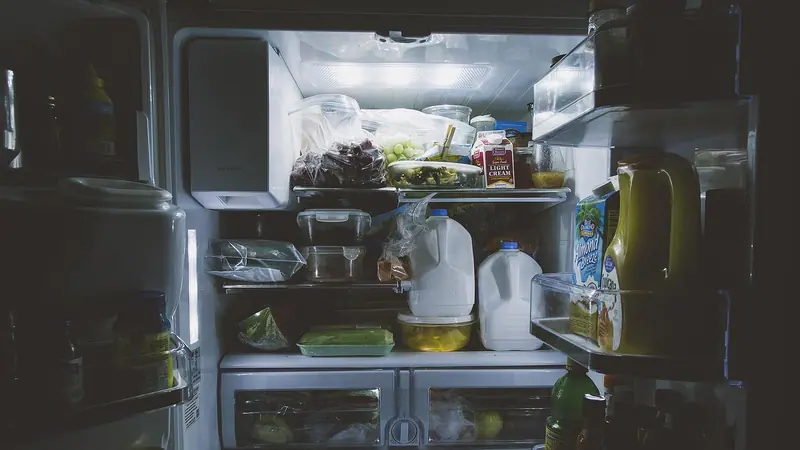 Geladeira aberta iluminando a cozinha escura, com prateleiras cheias de alimentos e garrafas