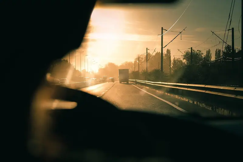 Vista de dentro de um carro em movimento, com o sol baixo ofuscando a estrada à frente