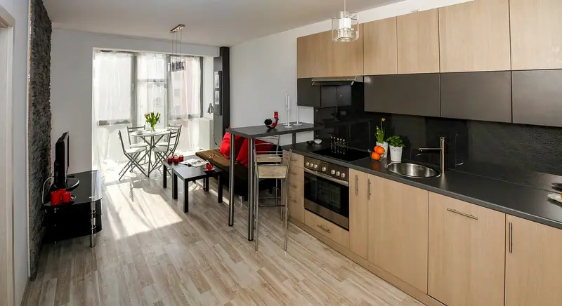 Apartamento compacto com cozinha encostada na parede, sofá, mesa alta e uma varanda envidraçada e clara ao fundo