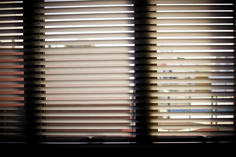 Persiana horizontal fechada diante de uma janela, com a luz do dia passando entre as palhetas