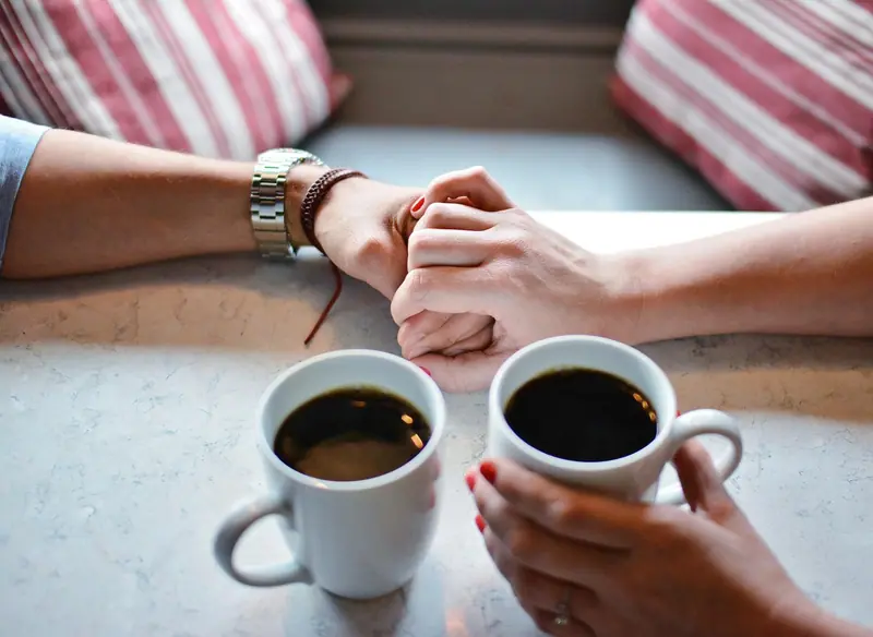 Duas pessoas à mesa, vistas só pelas mãos, cada uma com uma xícara de café