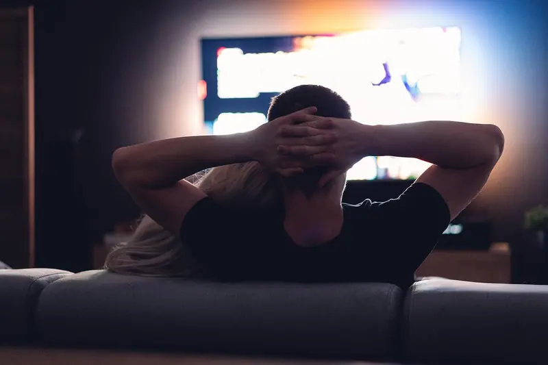 Pessoa de costas no sofá assistindo televisão numa sala escura