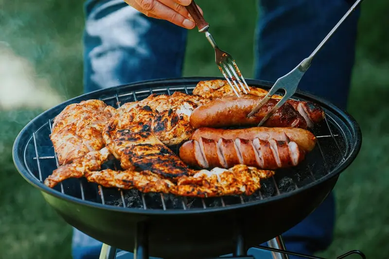 Mãos virando linguiças e carnes numa churrasqueira redonda